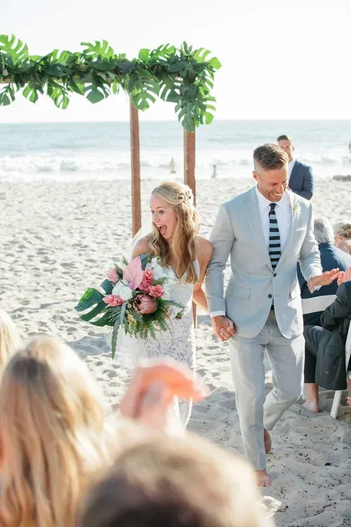 Bride and groom walking on a beach ceremony aisle under a floral arch