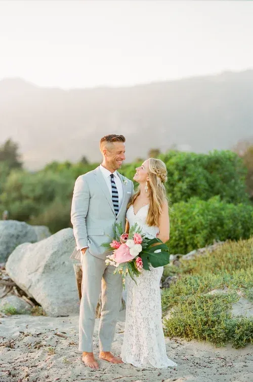 Couple standing outdoors in formal attire, holding a bouquet among rocks and greenery at sunset