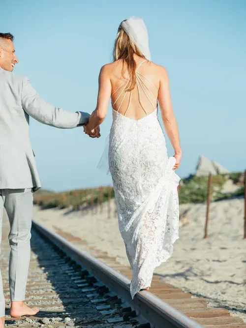 Couple walking barefoot along railroad tracks toward the beach, woman in white dress and hat