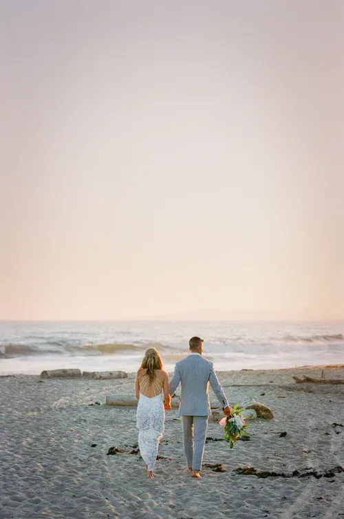 Couple walking barefoot on a beach at sunset, holding hands near flowers on the sand