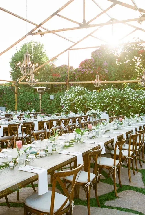 Outdoor dining setup with long white tables, wooden chairs, and greenery under a canopy at sunset