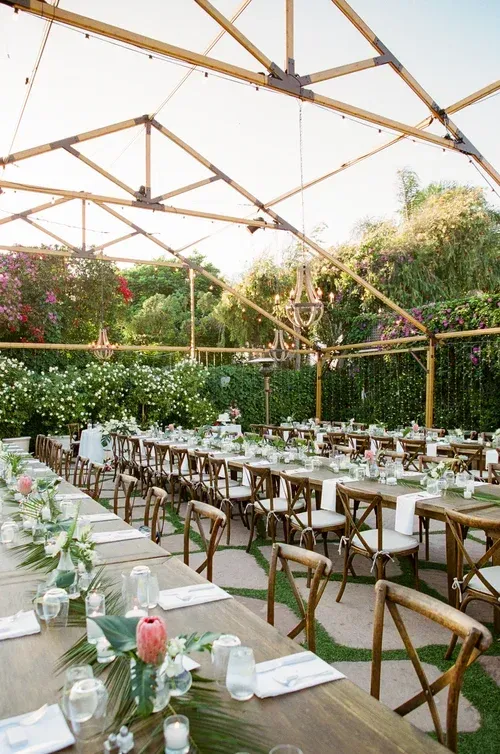 Outdoor garden dining setup with long white tables, wooden chairs, and greenery under a geometric canopy.