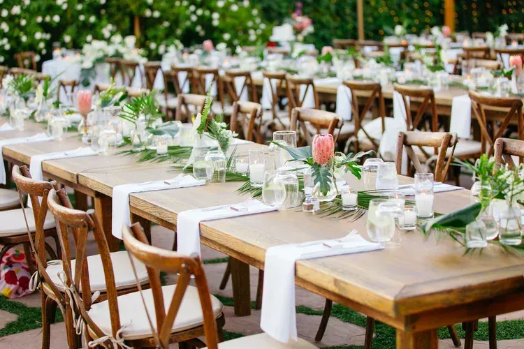Long outdoor banquet tables set with white runners, glassware, and greenery among wooden chairs in a garden