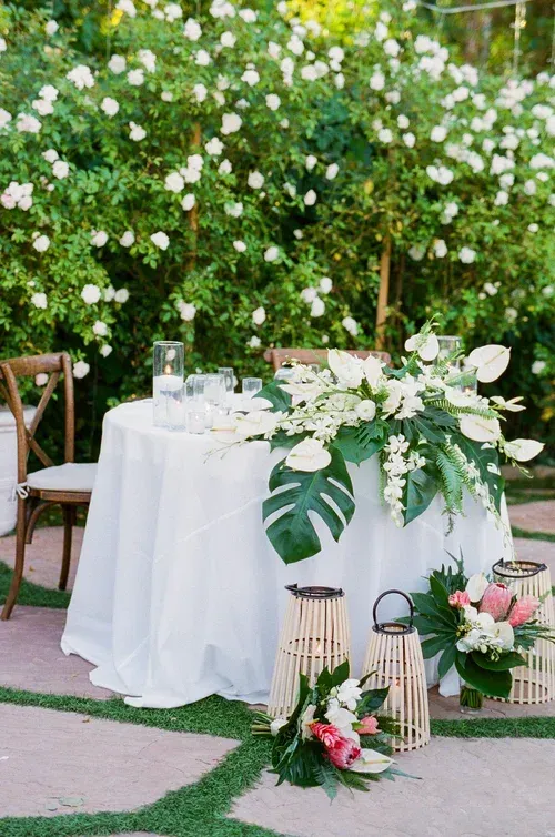 Outdoor wedding table with white tablecloth, white floral arrangements, lanterns, and greenery backdrop
