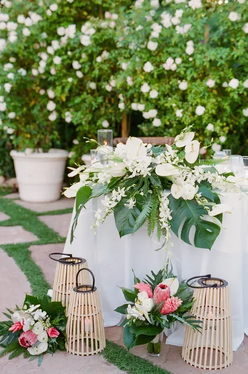 White floral wedding table display with greenery, pink bouquets, lanterns, and a lace-covered table outdoors