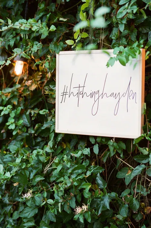 White square sign with handwritten hashtag text hanging on a leafy green wall beside a warm light