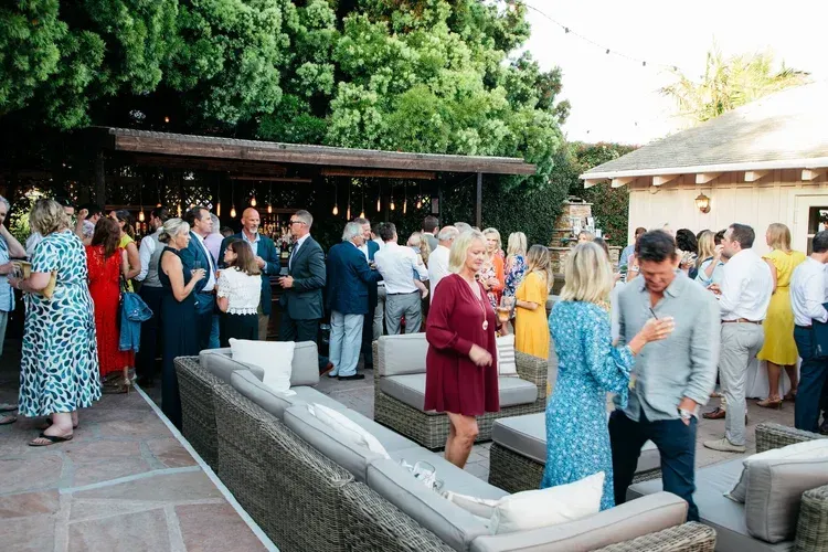 Outdoor party with guests mingling around a stone fire pit in a garden patio, several in colorful outfits