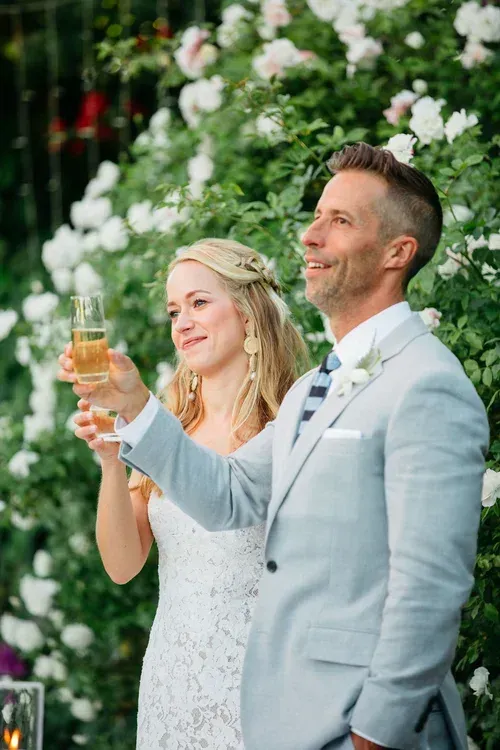 Bride and groom toast with champagne at an outdoor wedding, smiling in front of white flowers.