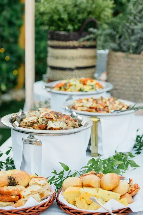 Buffet table with covered serving dishes and platters of food outdoors, decorated with greenery