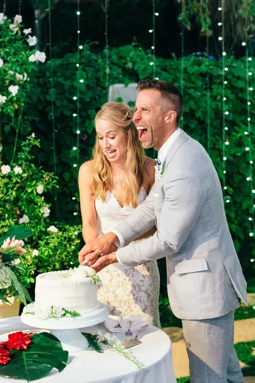 Couple in light outfits cutting a white wedding cake at an outdoor celebration with greenery and flowers