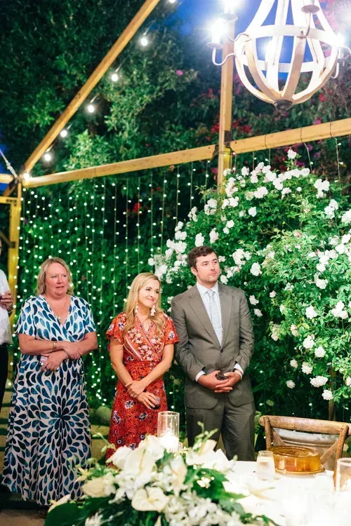 Three guests stand in a decorated garden venue with greenery, white flowers, and string lights.