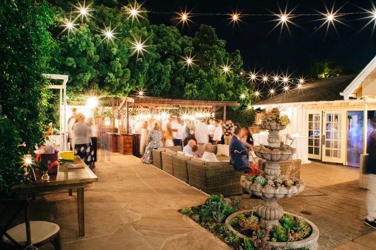Outdoor evening gathering with string lights, people mingling around tables, and a tiered fountain in the foreground