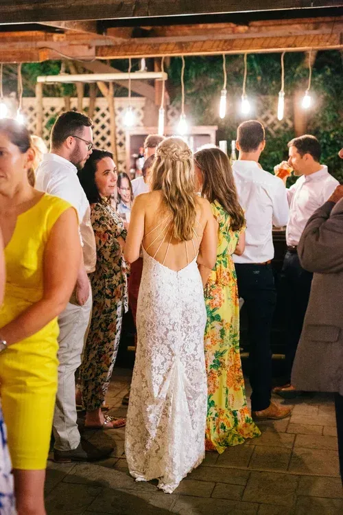 Guests mingling at an outdoor evening party, with string lights and a woman in a white lace dress centered.