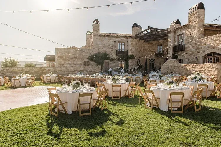 Outdoor wedding reception with round tables and white chairs on a lawn beside a stone building at sunset