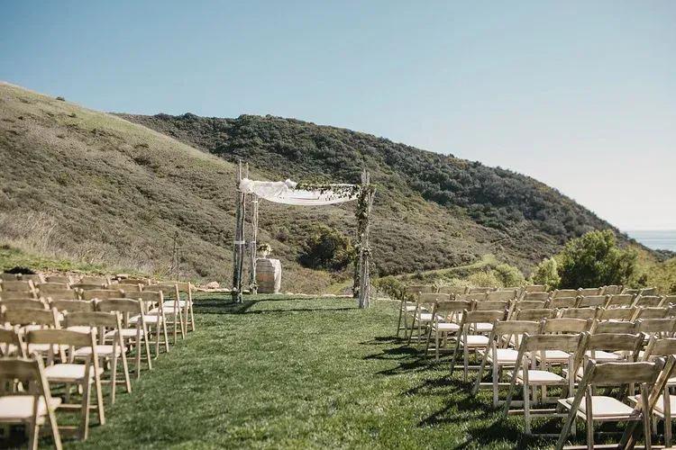 Outdoor wedding ceremony setup on a grassy aisle between rows of chairs, framed by hills and a white arch.