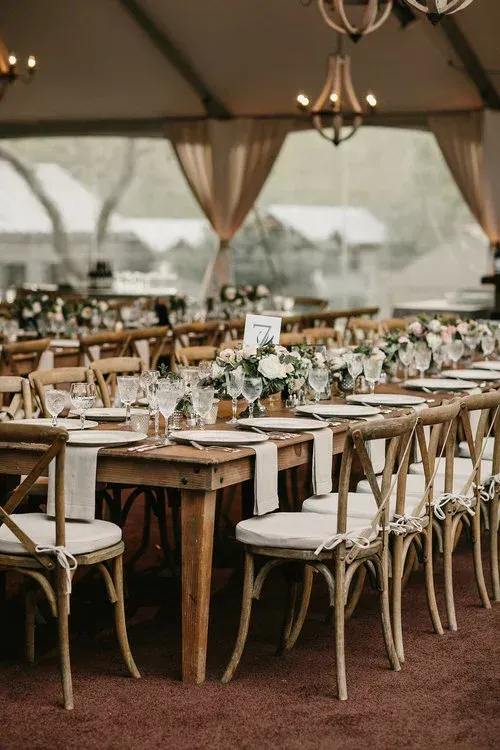 Decorated outdoor banquet tables under a tent, set with white chairs, flowers, and candles.