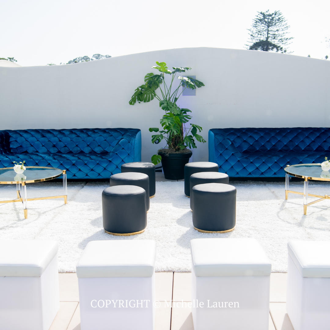 Modern outdoor patio with blue sofas, gray ottomans, white seating, and a potted tree under bright sun