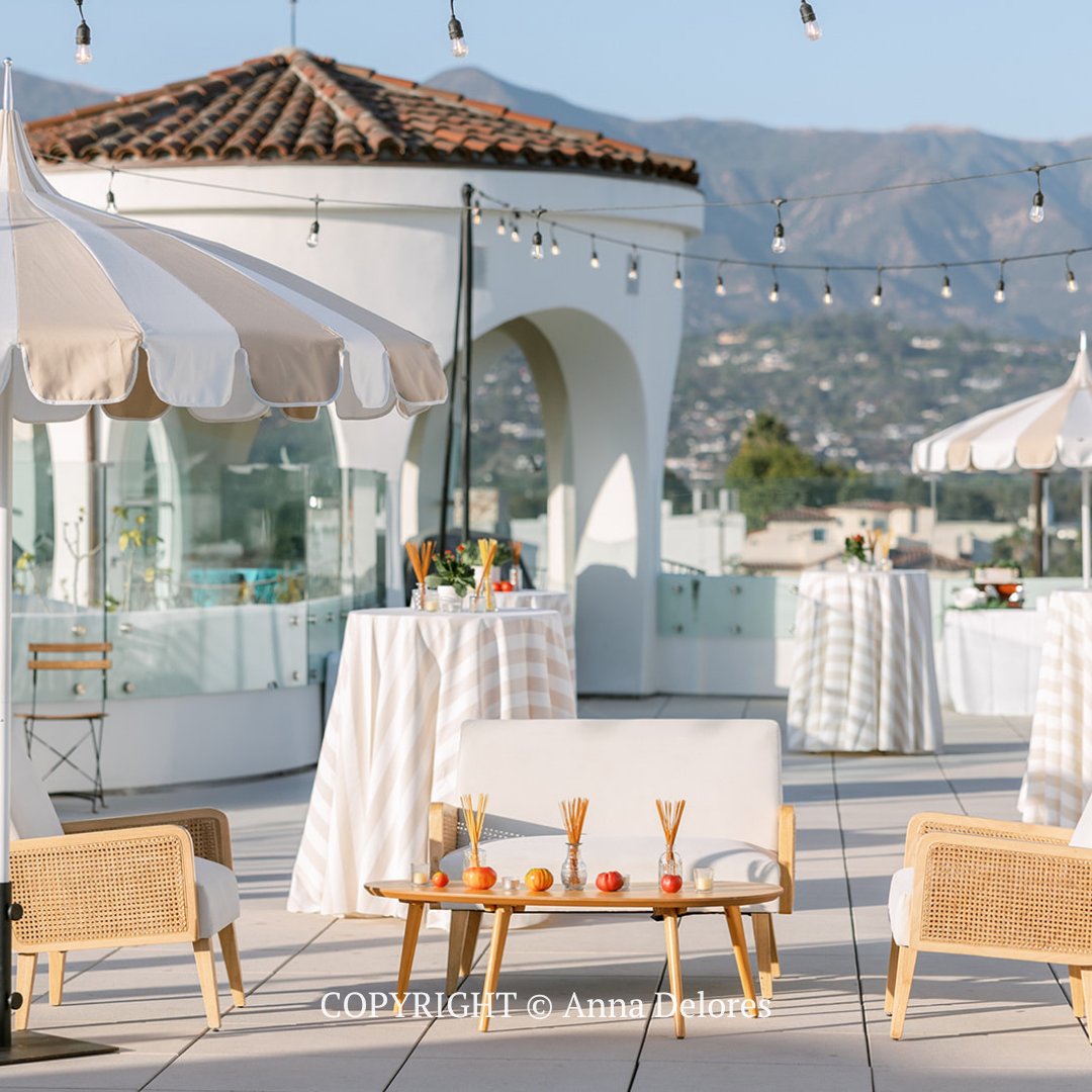 Outdoor café terrace with white tables, umbrellas, string lights, and mountain views under a blue sky