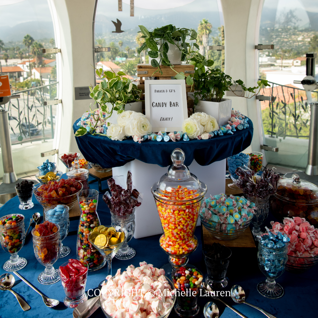Candy buffet on a decorated table with glass jars and a sign in a bright arched room with city views
