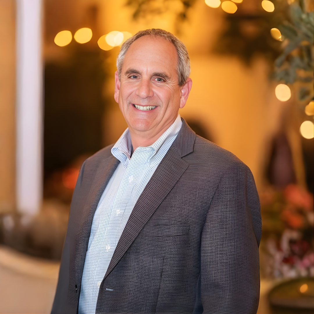 Smiling man in a striped blazer standing in a warmly lit indoor setting with blurred lights in the background