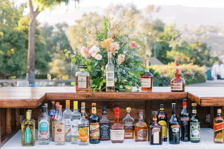 Outdoor bar table lined with assorted liquor bottles and floral arrangements in a sunny garden setting