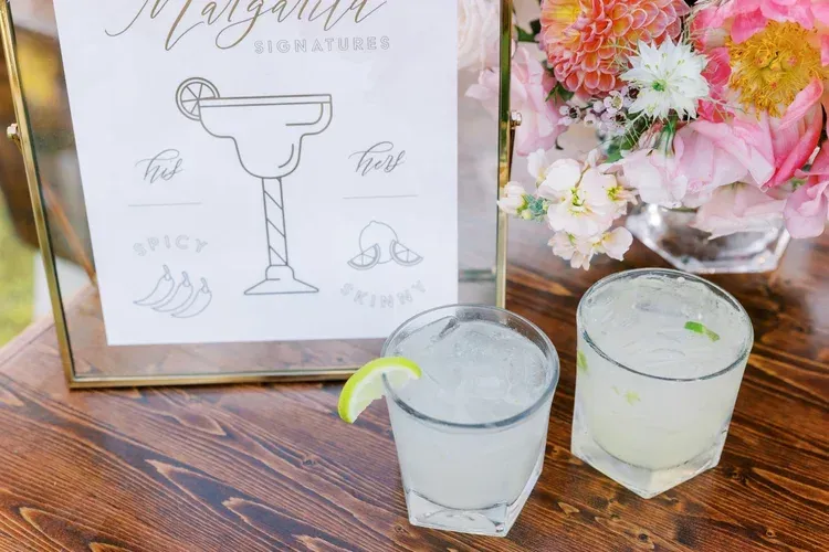 Framed margarita sign beside two frosty lime drinks on a wooden table with pink flowers.