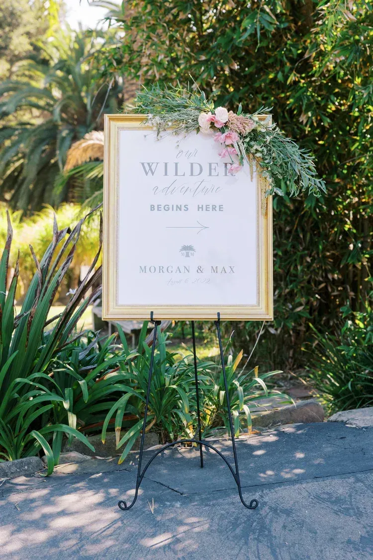 Floral wedding welcome sign on an easel in a garden with greenery and palm trees