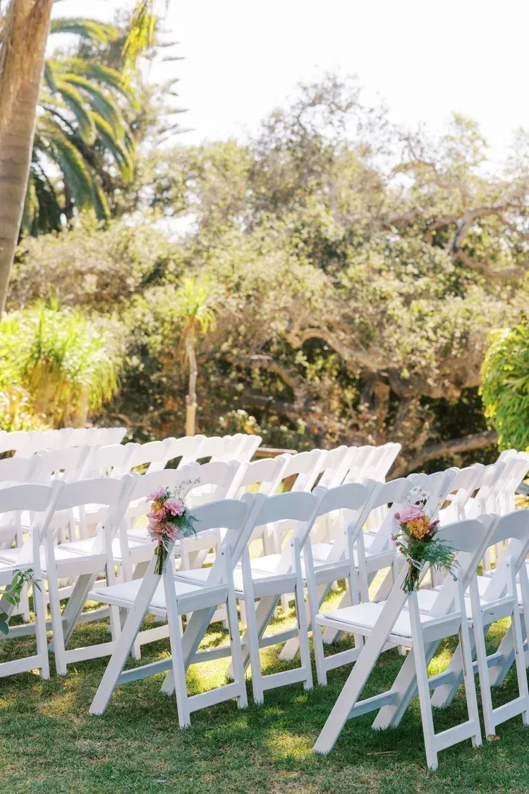 Rows of white chairs on grass with small floral bouquets, set outdoors in sunlight.