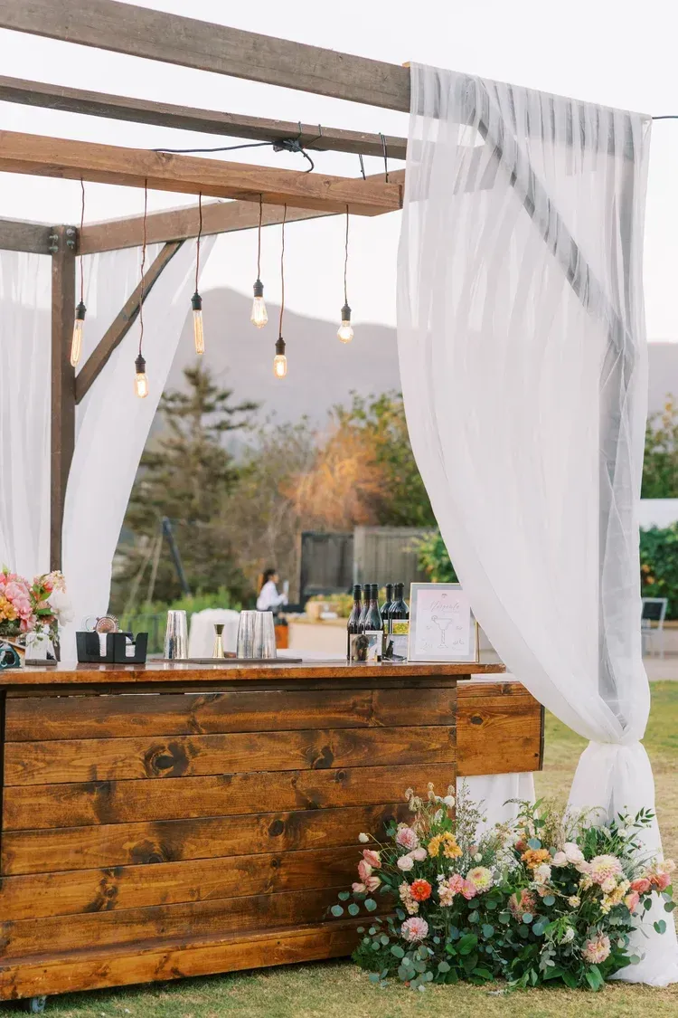 Rustic outdoor wedding bar with white drapes, hanging lights, and floral arrangements under a pergola