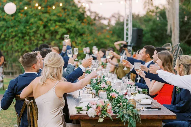 Guests toast at a long outdoor dinner table with flowers and candles.