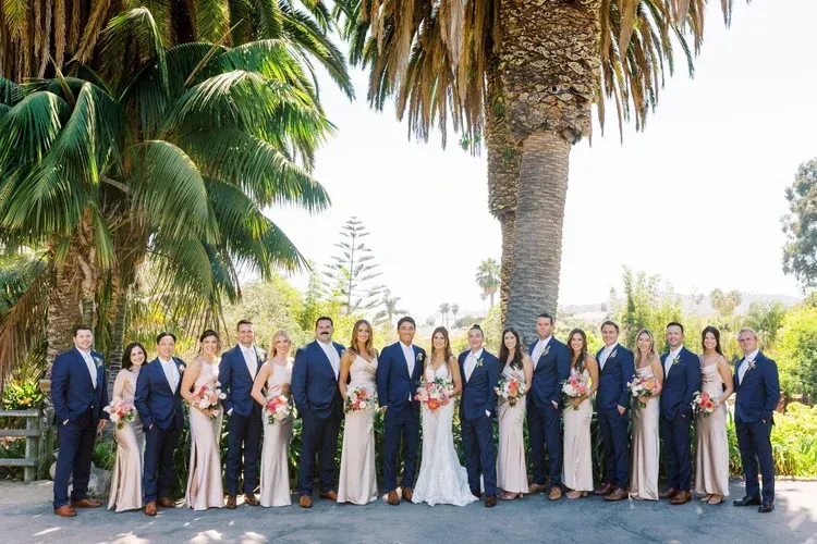 Large wedding party posing outdoors beneath palm trees, with bridesmaids in navy and white dresses
