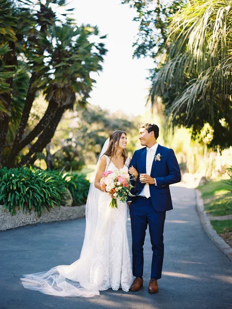 Bride and groom walking arm in arm on a sunny garden path, smiling and holding a bouquet