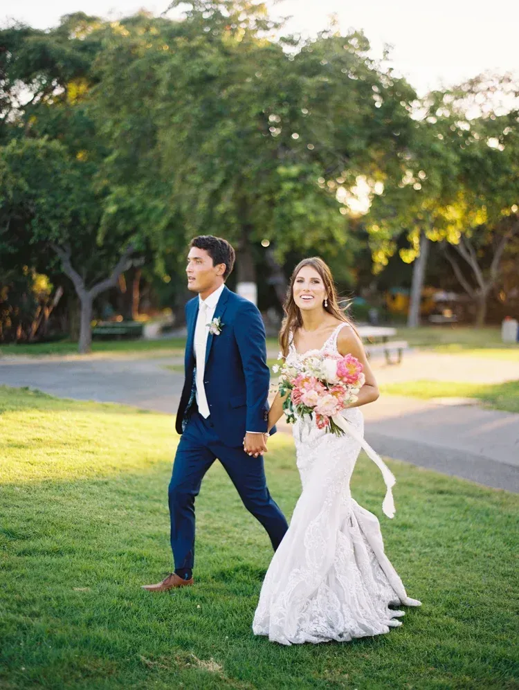 Bride and groom walking hand in hand on a grassy lawn in formal wedding attire at sunset.