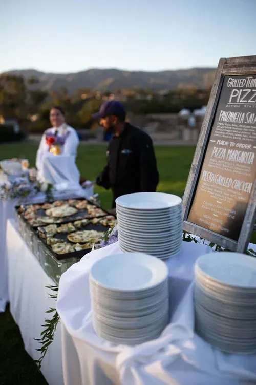 Outdoor buffet table with stacked white plates, trays of food, and a menu sign at sunset