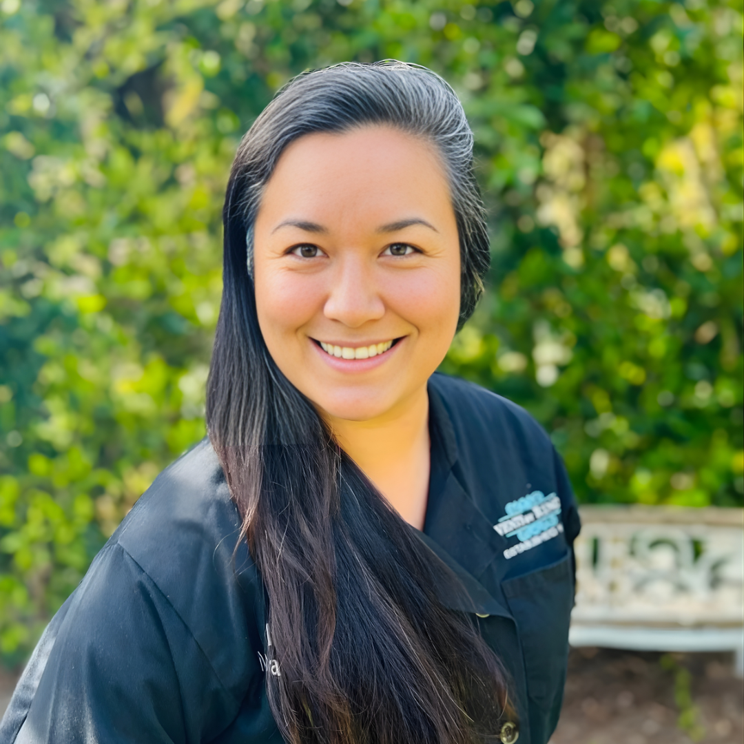 Smiling person in a black top outdoors with green foliage and a bench in the background