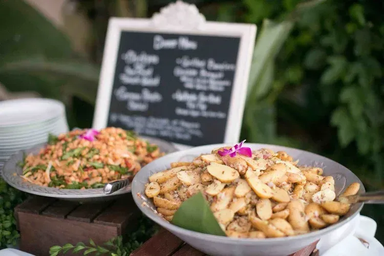 Buffet dishes of pasta and salad in front of a black menu board at an outdoor event