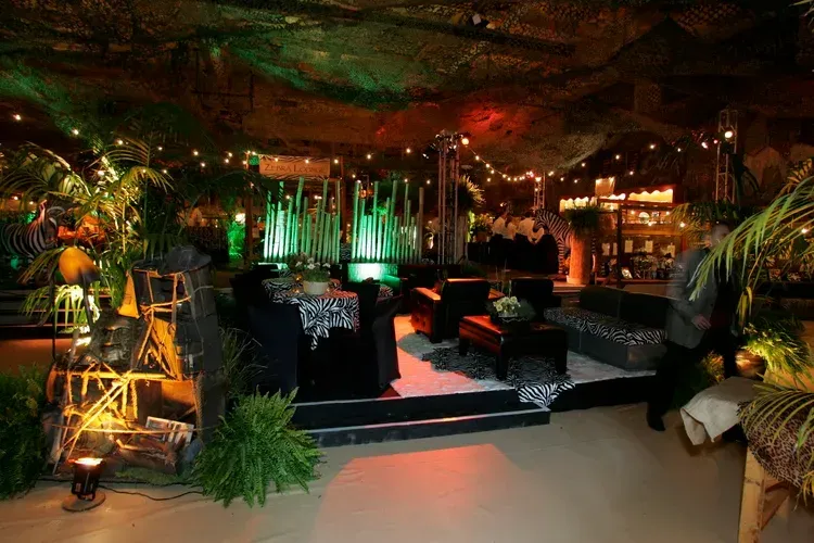 Dimly lit tropical lounge with green accent lighting, plants, and black seating on a stage-like platform
