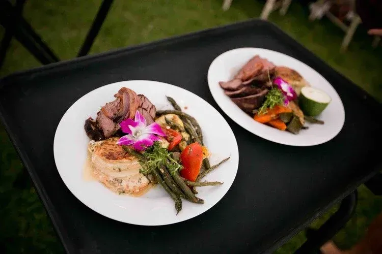 Two plated gourmet meat dishes with vegetables on a black table outdoors
