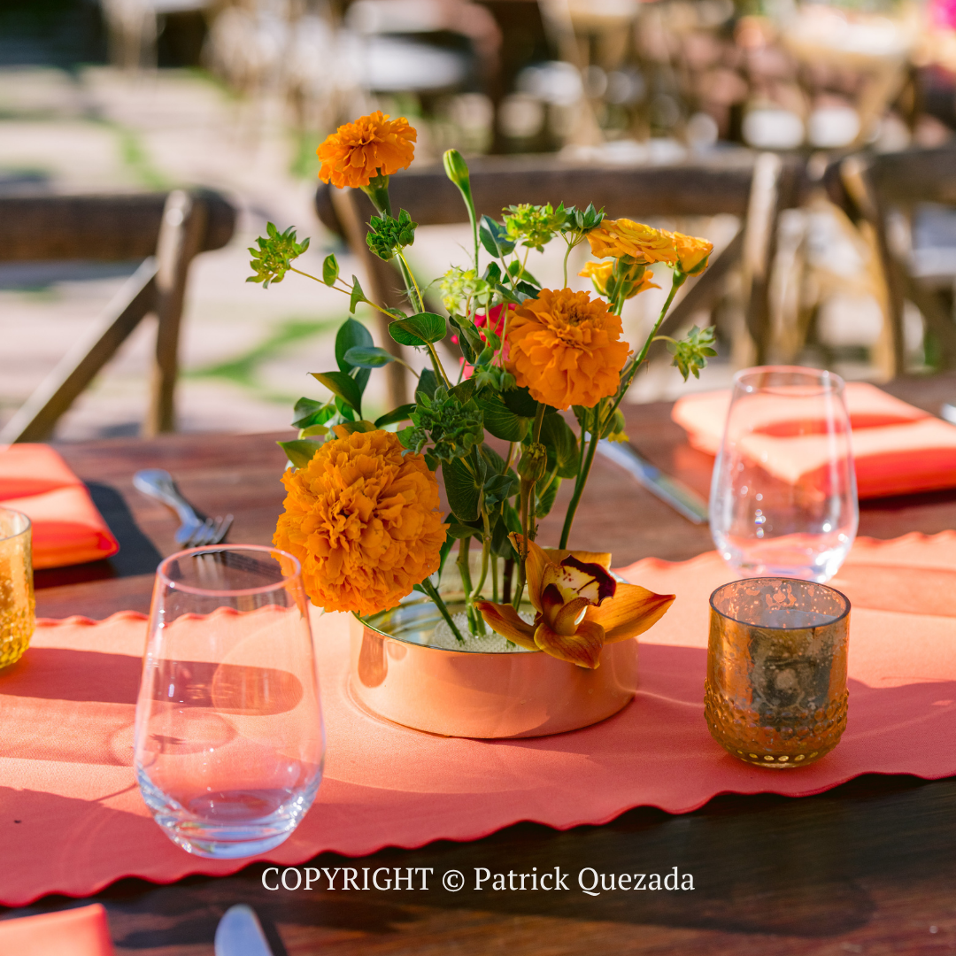 Orange flowers in a bronze pot on a pink tablecloth with wine glasses outdoors