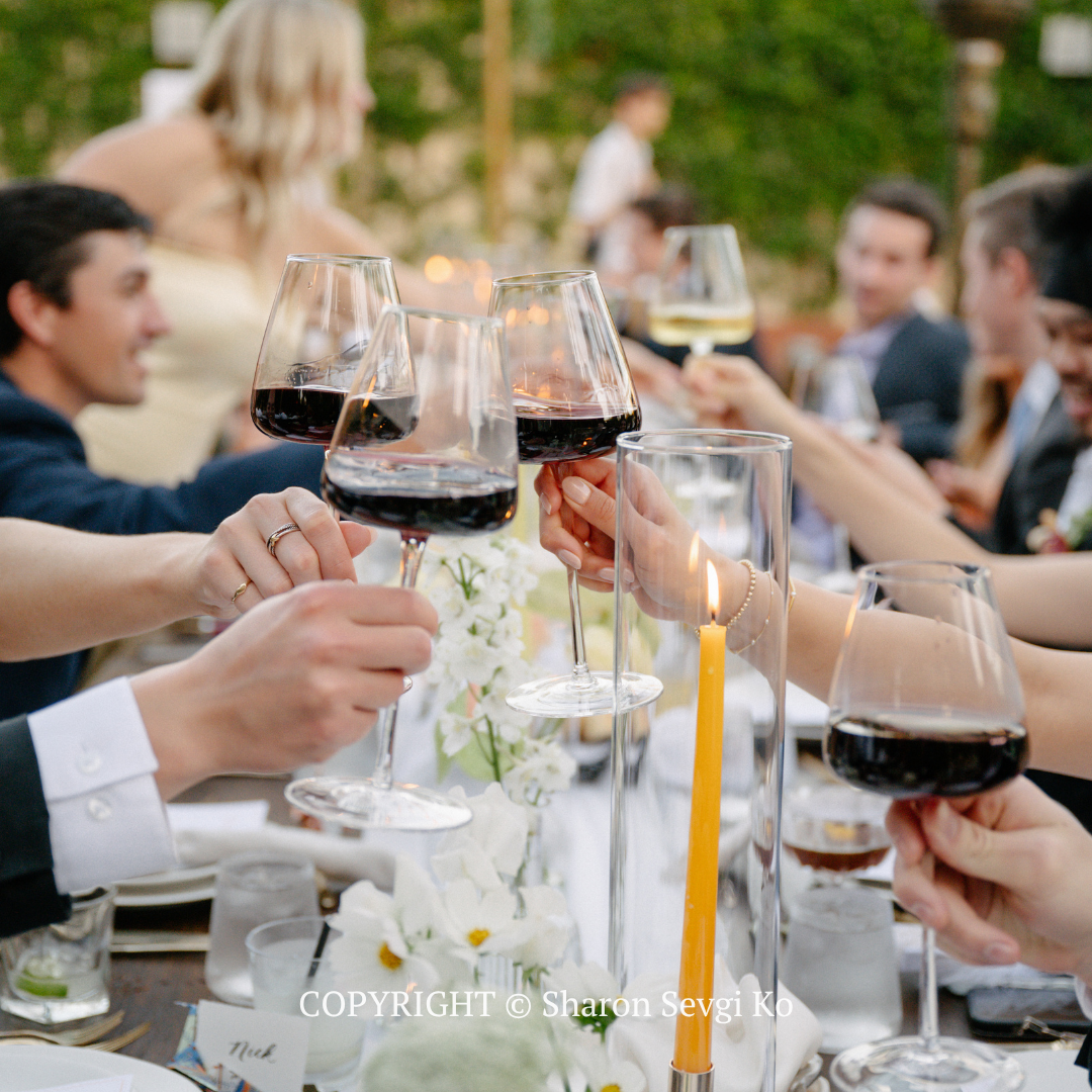 People clinking wine glasses at an outdoor dinner party with candles on the table