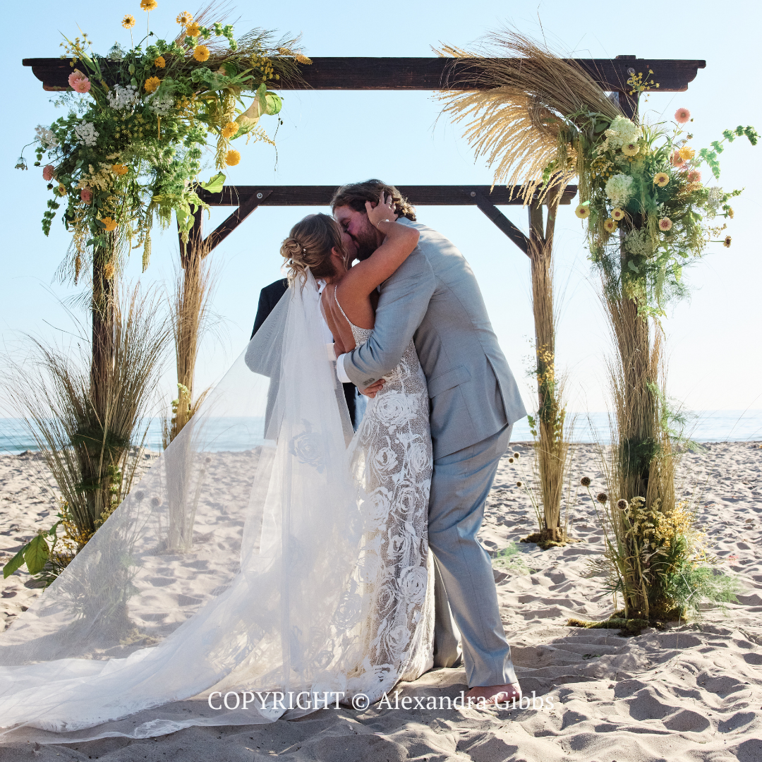 Couple embracing under a floral arch on a sandy beach at a wedding ceremony