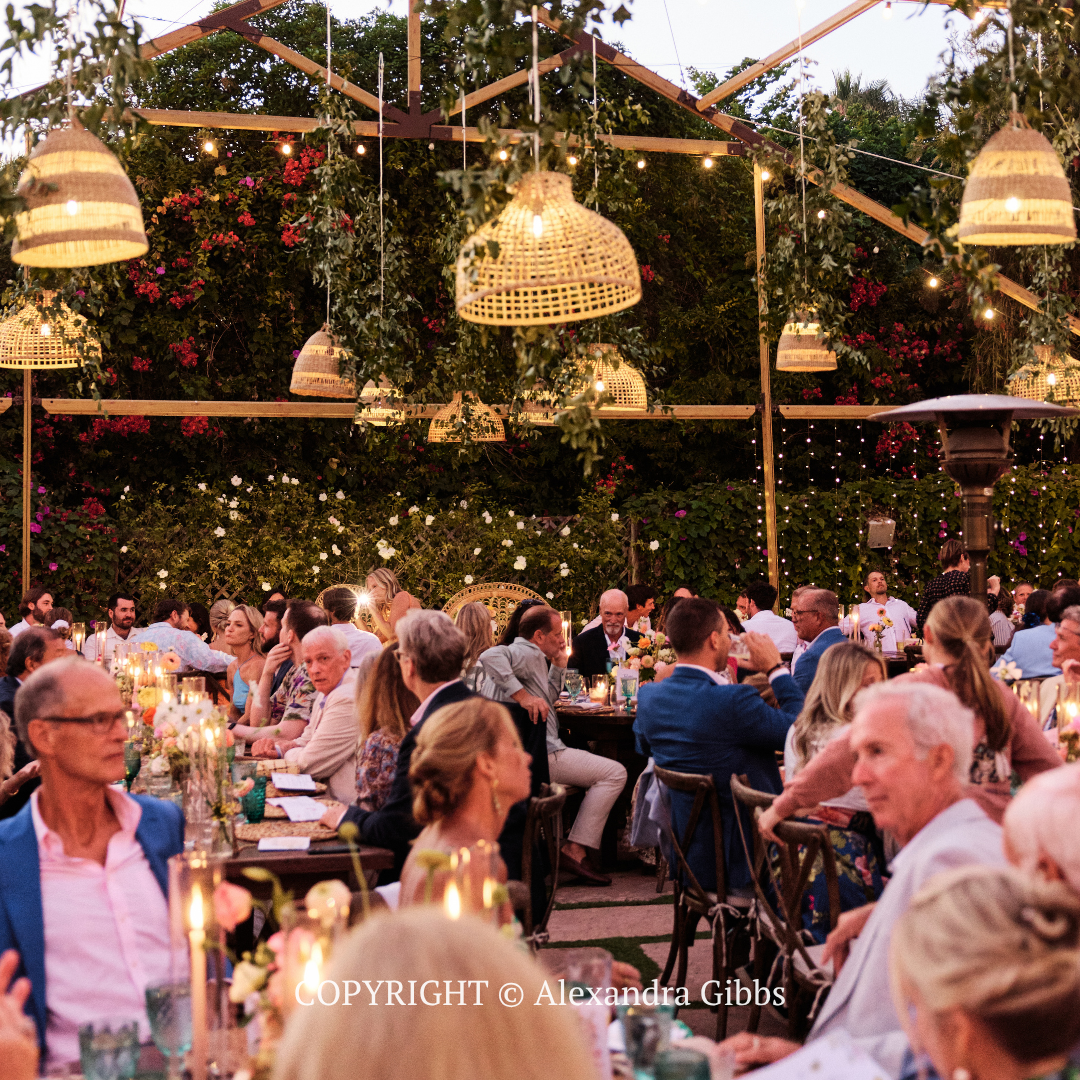 Crowded outdoor dinner party under hanging lights in a garden at dusk