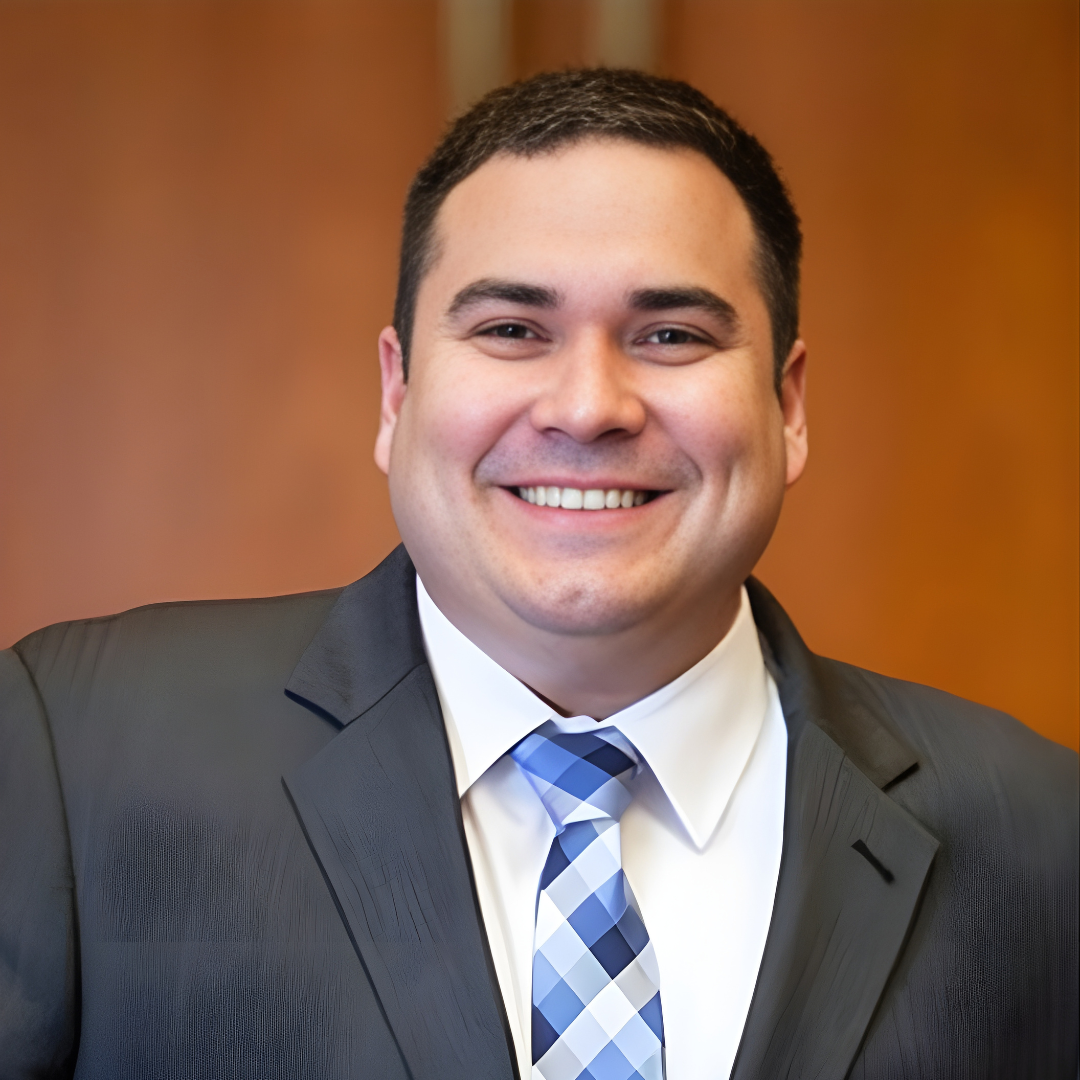 Smiling man in a dark suit and blue plaid tie against a warm blurred background