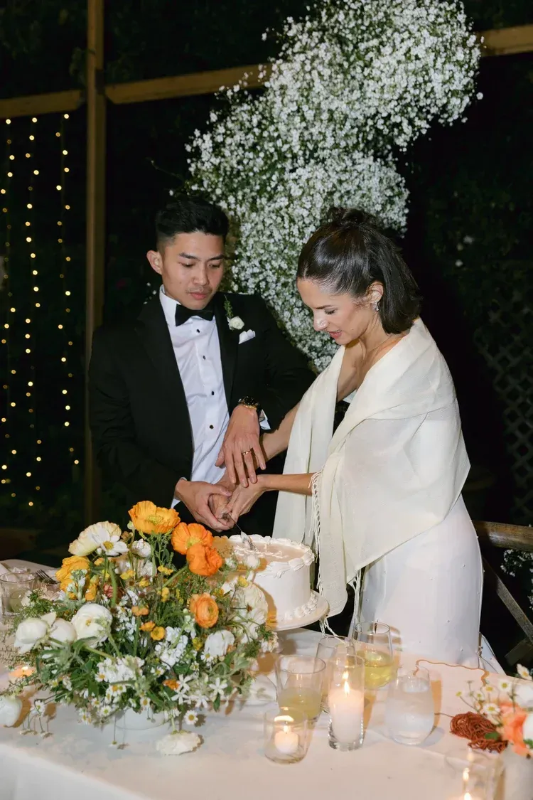 Couple cutting a cake at a candlelit wedding table with white flowers and orange accents