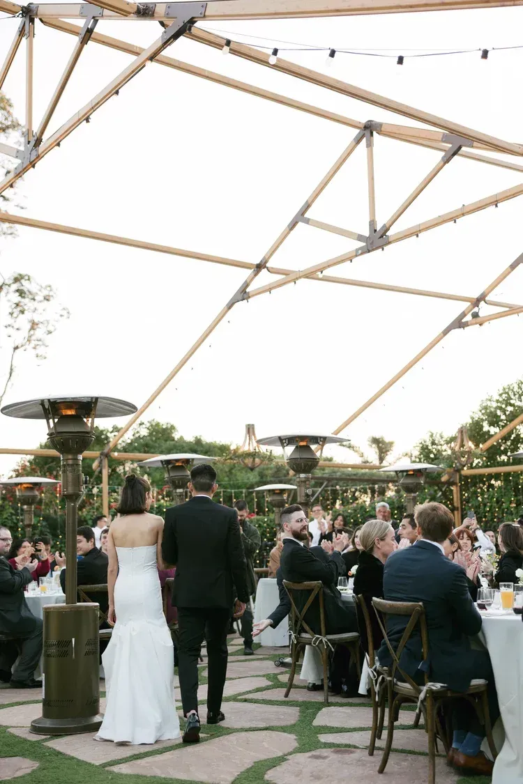 Outdoor wedding ceremony with bride and groom walking beneath a white canopy, guests seated nearby