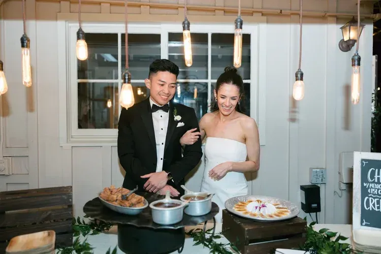Bride and groom serving food at a buffet table under hanging lights