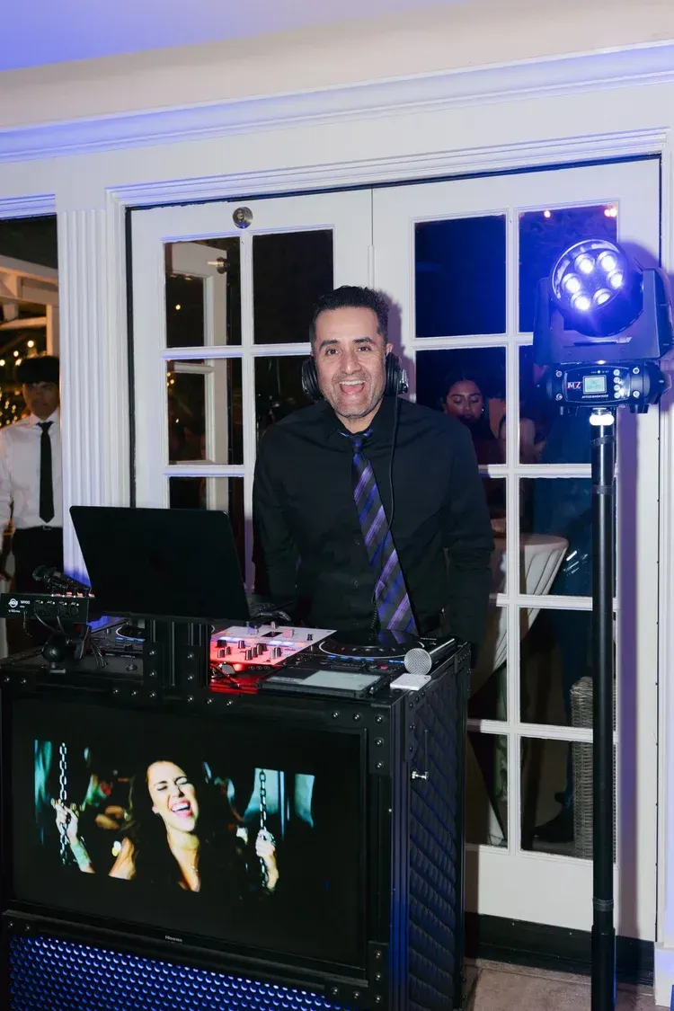 Man standing behind a DJ booth with glowing blue lights in a room with white French doors.