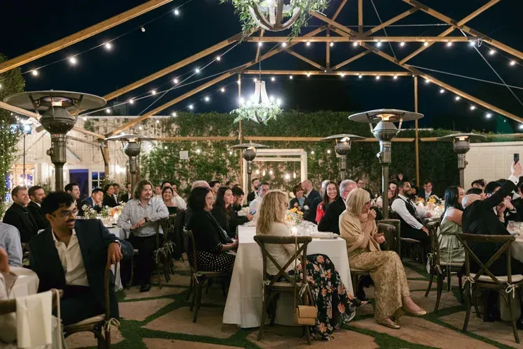 Guests seated under string lights at an outdoor evening event, with a speaker at the front.