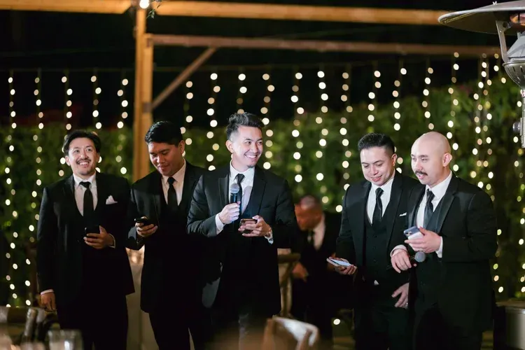 Five men in dark suits smiling and holding drinks at an outdoor evening event with string lights