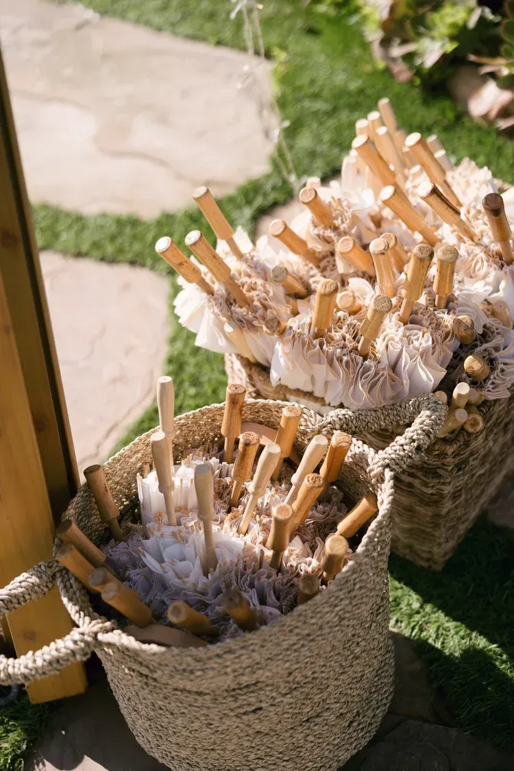 Woven baskets filled with white candles wrapped in orange paper, displayed outdoors on grass and paving stones